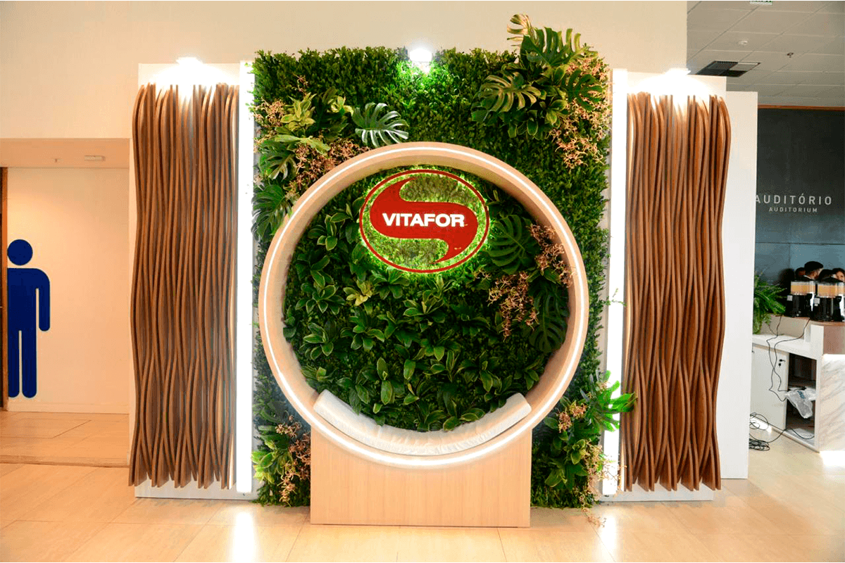 Painel verde com vegetação, logotipo central, aro circular iluminado e laterais de ripas curvas amadeiradas.