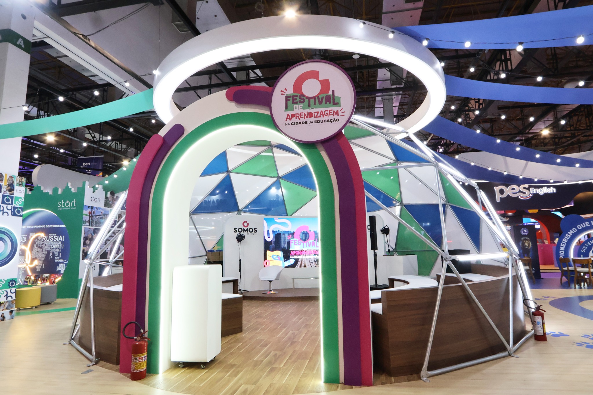 Estande com cúpula de painéis triangulares, arco colorido iluminado, testeira circular branca e área interna de apresentação.