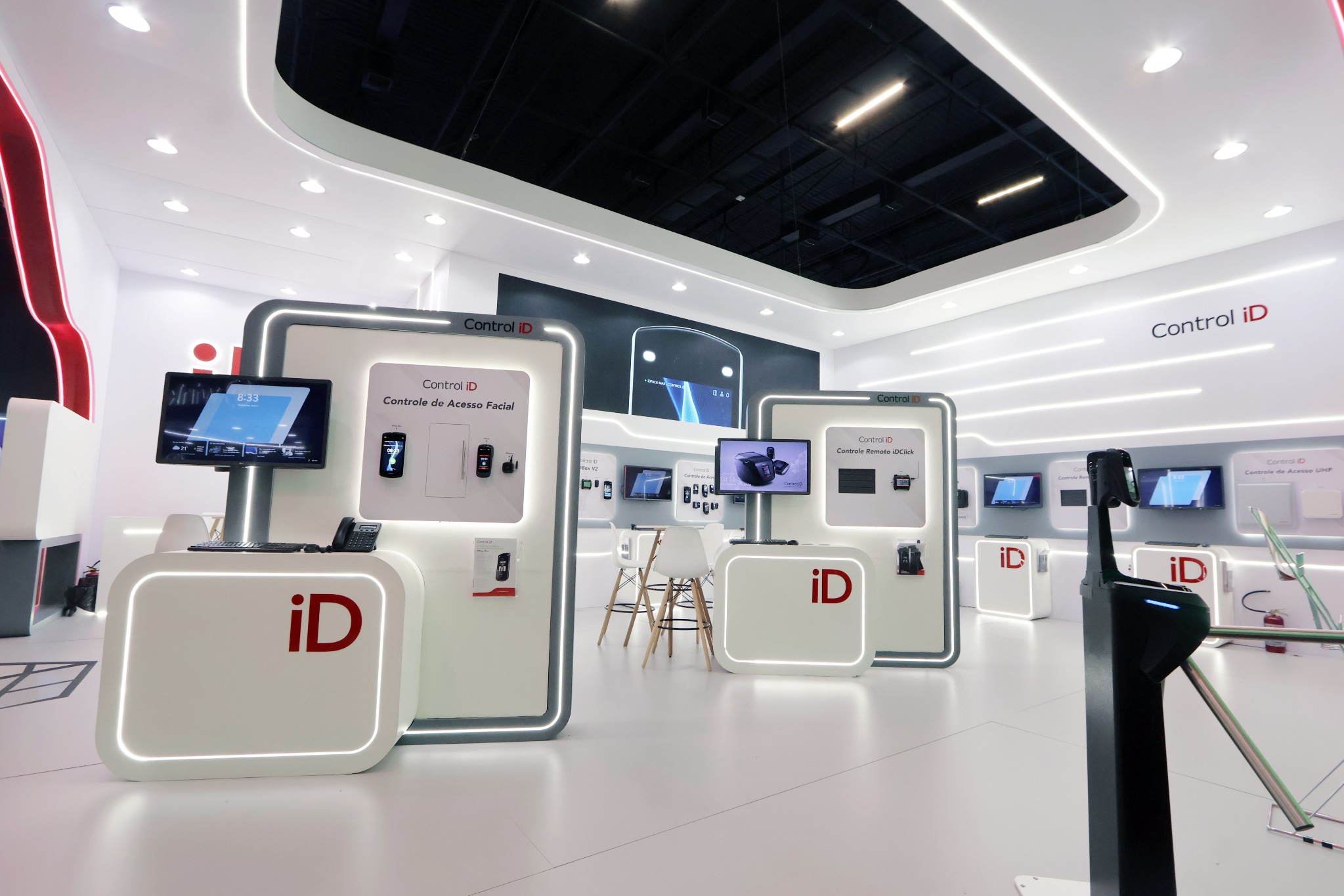 Interior de estande branco com expositores de produtos, monitores, balcões com contornos iluminados e testeira curva.
