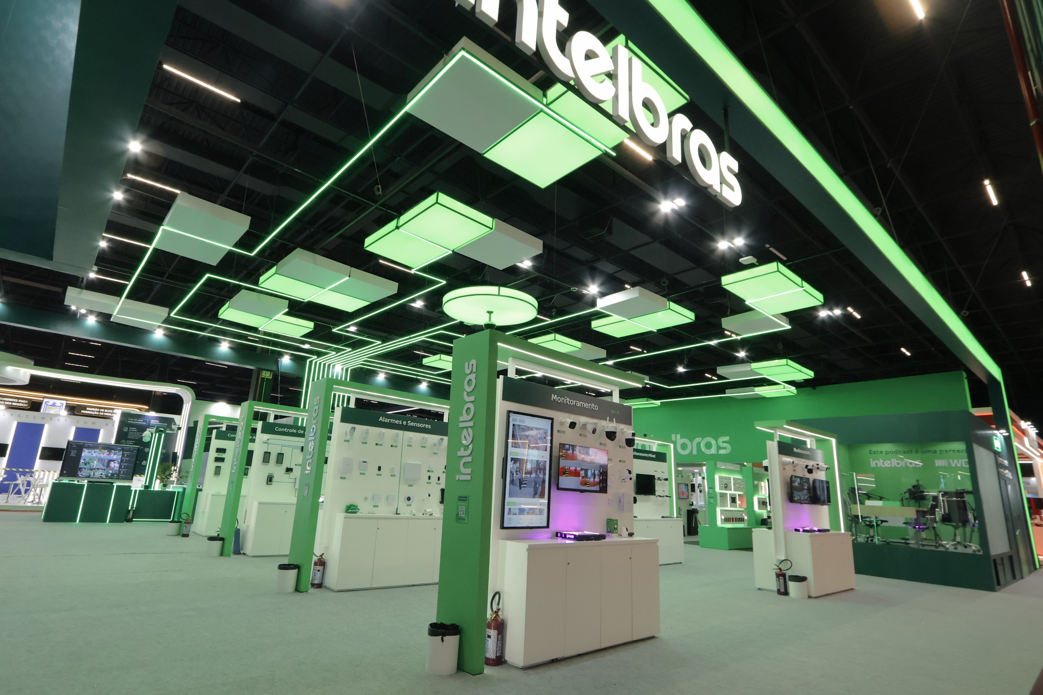 Estande verde e branco com painéis de demonstração de equipamentos, iluminação linear e elementos geométricos suspensos.
