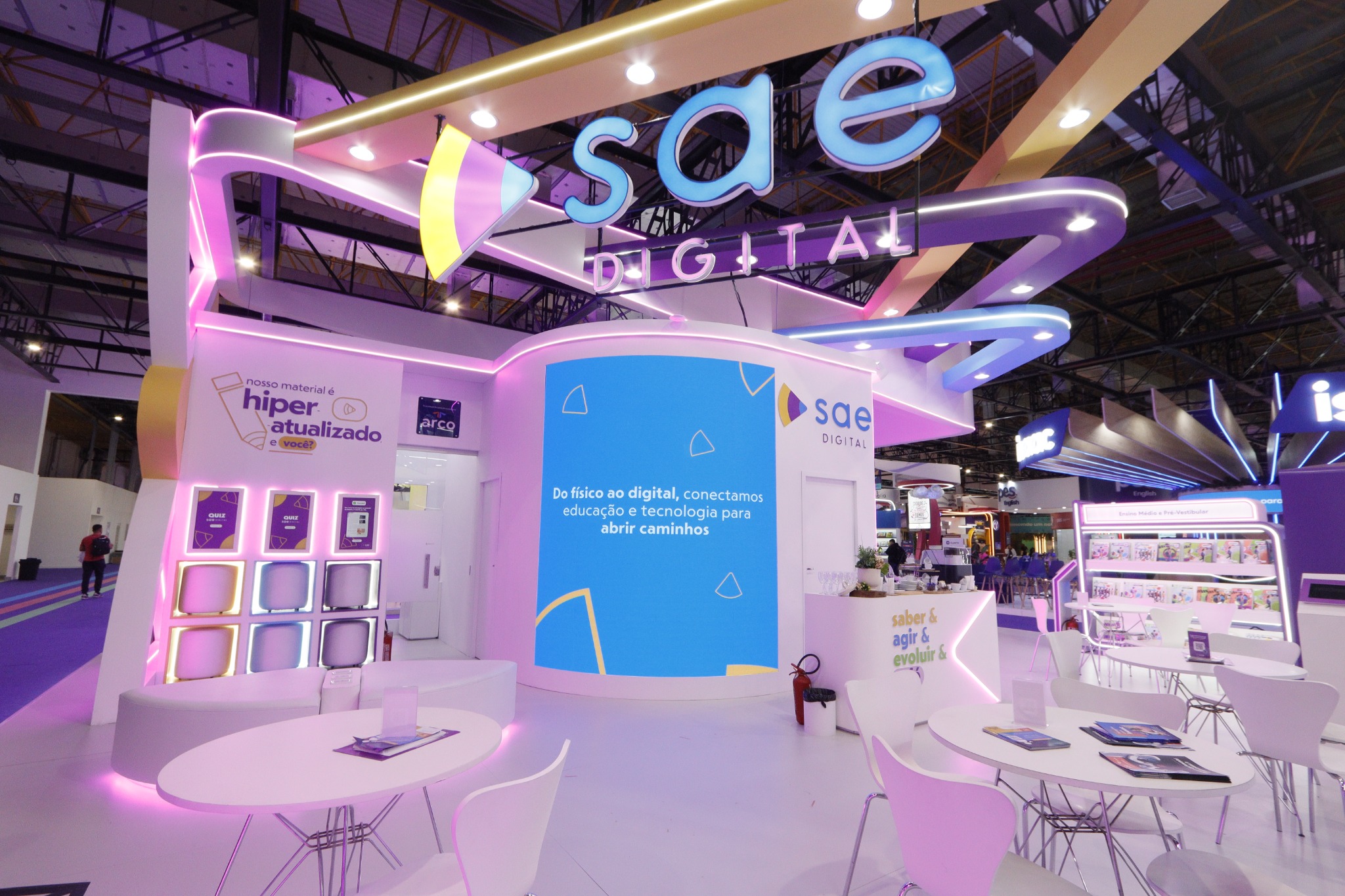 Estande da SAE Digital com painel central azul, testeiras curvas iluminadas, expositores de livros e mesas de atendimento.