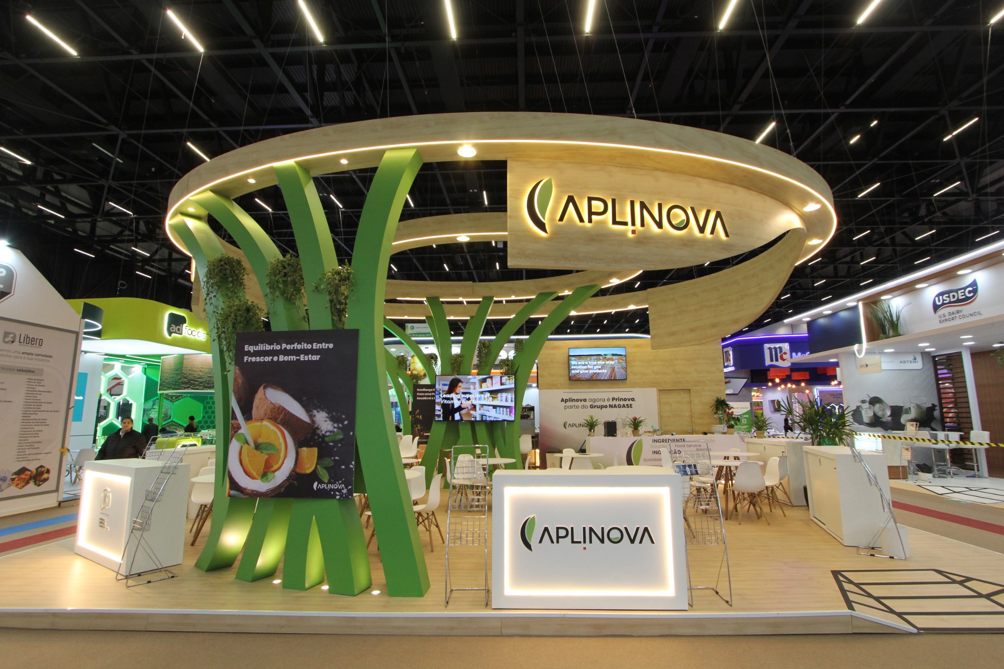 Estande da Aplinova com testeira circular amadeirada, estruturas verdes, vegetação suspensa e balcões brancos iluminados.