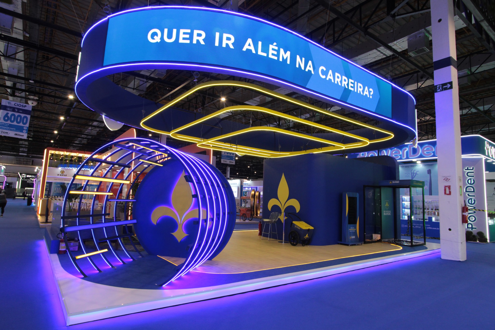 Estande azul com testeira curva suspensa, painel digital, estrutura circular vazada e iluminação violeta e amarela.